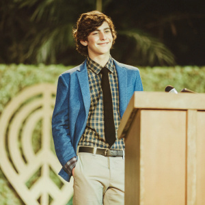 David Mazouz, young actor, wearing a plaid shirt, black tie, and blue blazer. Standing confidently against a dark blue backdrop adorned with a large, logo-like graphic design. Event photo.