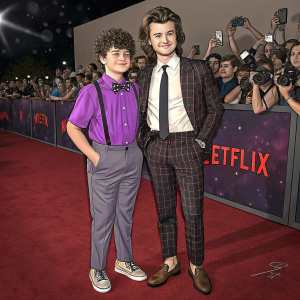 Gaten Matarazzo and Joe Keery at a Netflix event. Gaten in a vibrant purple shirt and casual shoes. Joe in a sharp white shirt, dark tie, and plaid suit with loafers. Background filled with fans and cameras.