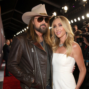 Billy Ray Cyrus, in a stylish brown leather jacket, sunglasses, and a cowboy hat, alongside Tish Cyrus in a chic white dress. The duo is at a glitzy award show with flashing cameras and a glamorous audience.