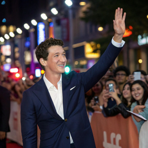 Miles Teller, dressed in a navy suit and white shirt, smiling and waving to the crowd at a film festival. Crowded outdoor setting, vibrant lights, and enthusiastic fans.
