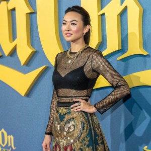 Jessica Henwick attends the 'Glass Onion: A Knives Out Mystery' premiere at the London Film Festival. Dressed in a black mesh top and an elaborate gold-embellished skirt. The background is a bold yellow and blue design with text.