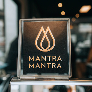 The Mantra Manta logo features a radiant golden flame symbol set against a sleek black backdrop. Below the logo, the name 'Mantra Manta' is elegantly scripted in bold, modern font, shimmering with gold against the dark background.