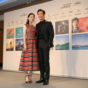 Lee Na-Young and Daniel Henney at the Cannes Film Festival press conference. Lee Na-Young in a vibrant striped dress with sleek black heels. Daniel Henney in a sleek black suit. Behind them, a backdrop of movie posters.