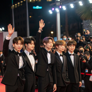 Yeonjun, Soobin, Huening Kai, Taehyun, and Beomgyu from Tomorrow X Together at a glitzy awards ceremony. Wearing tuxedos, they wave at the audience. Flashing lights and paparazzi in the background.