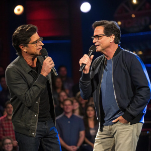 John Stamos and Bob Saget performing on the Late Show with Stephen Colbert stage. Both wear casual outfits. The audience is blurred.