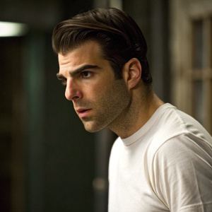 Close-up of Zachary Quinto, dressed in a white t-shirt, intense expression. Dark, blurred backdrop.