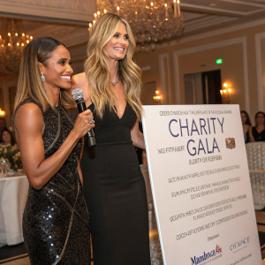 Ciara and Heidi Klum at a charity gala. They are standing beside a giant banner promoting the event. Heidi is using a microphone and Ciara is smiling