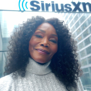 Angela Bassett, mature actress with a warm smile, in a close-up portrait. She wears a white, sequined turtleneck. SiriuxXM logo backdrop. Professional setting.