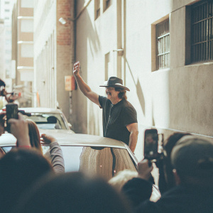Robert Rodriguez at a bustling city street, promoting 'Machete'. Wearing a black t-shirt and hat. Surrounded by fans and cameras.