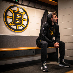 Patrice Bergeron, wearing a black hoodie and sweatpants, sits on a bench in a locker room with Boston Bruins logo behind him.