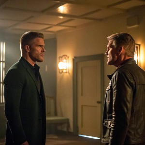 Stephen Amell as Oliver Queen and Dolph Lundgren as Anatoly Kuragin at a tense meeting scene in Arrow. They stand in a dimly lit room with soft, warm lighting
