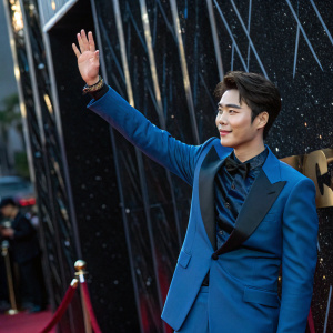 Li Yifeng, Chinese actor and singer, poses elegantly at a Hollywood award show. Wearing a blue suit, he waves to the audience. The backdrop is a shiny black surface with subtle reflections.