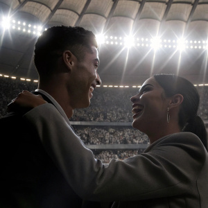 Cristiano Ronaldo and Georgina Rodriguez laughing and hugging. Brightly lit indoor stadium. Crowds of fans in the background.