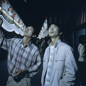 Jordan Dang and Mikey Kawata, as Young Ando and Young Hiro respectively, in an animated scene. Hiro, an Asian male, looks intensely at something while wearing a plaid shirt. Ando, an Asian male, is in a light jacket, expression neutral. They are in a bustling area filled with vibrant decorations and people.