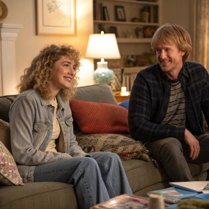 Imogen Poots and Owen Wilson in a cozy living room scene from 'She's Funny That Way'. Curly hair, casual attire. Warm lighting, soft furnishings
