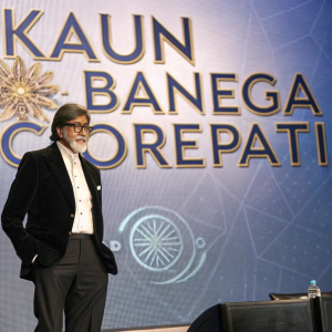 Amitabh Bachchan at a press conference. Wearing formal attire with a black suit and white shirt. Standing beside a large screen displaying the 'Kaun Banega Crorepati' logo.