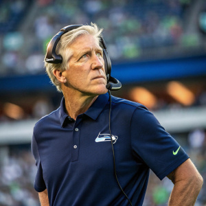 Pete Carroll, head coach of the Seattle Seahawks, wearing a navy blue team polo shirt. Serious expression. Wearing a headset. Outdoor stadium background.
