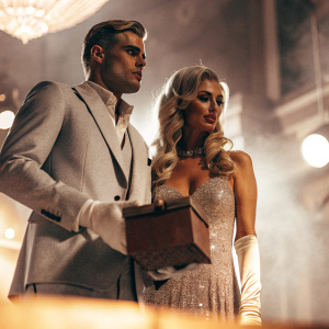 Paris Hilton and River Viiperi at an exclusive night gala. Paris in a sparkling evening gown, holding a mysterious box. River in a sophisticated gray suit. Both are standing on a dimly lit stage under soft, atmospheric lighting.