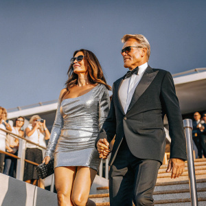 Jenny Sandersson and Dolph Lundgren at the Cannes Film Festival. She's in a sleek silver mini-dress, he's in a black tuxedo. Flashing lights and excited crowd in background.