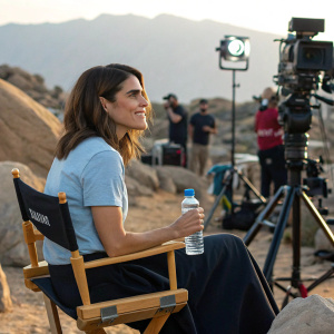 Karla Souza, seated outdoors in a director's chair amidst a rocky landscape, holding a water bottle. Wearing a casual light blue t-shirt and dark skirt. Surrounded by film crew lighting equipment.