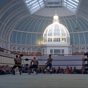 Lucha Britannia match at the Great British Tattoo Show. Indoor arena, ornate building backdrop. Wrestlers from diverse ethnic backgrounds in action. Audience focused on the ring.