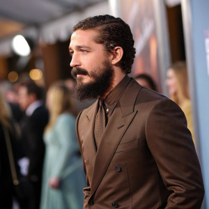 Shia LaBeouf, with a bushy beard, dressed in a brown suit. Standing at a Hollywood party with blurred guests in the background.