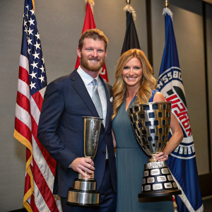 Dale Earnhardt Jr. and Amy Reimann at the NASCAR Hall of Fame induction. Posing with racing trophies, surrounded by team flags.