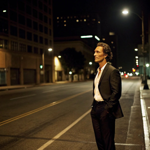 Matthew McConaughey in a candid shot, wearing a tailored black suit and white shirt, standing alone on a deserted city street at night.