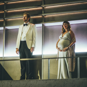 Carter Hudson and Hollye Gilbert at the premiere of Snowfall. She is pregnant. They stand against a gradient purple backdrop.
