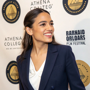 Alexandria Ocasio-Cortez, a young Latina politician, posing with a warm smile. She wears a navy blue blazer over a white blouse. The backdrop features the Barnard College and Athena Film Festival logos in gold and black.