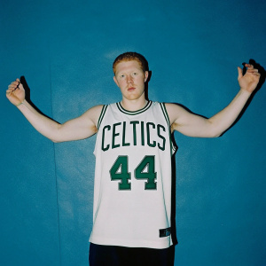 Brian Scalabrine, a former NBA player, in a Celtics jersey. White jersey with green 'Celtics' and number '44'. Standing against a solid blue backdrop. Determined expression, strong jawline.