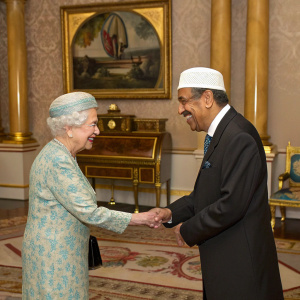 Queen Elizabeth II and Dr. Akbar S. Ahmed in a warm handshake moment. Inside the grand, opulent hall of Buckingham Palace, adorned with intricate gold detailing, an ornate wooden desk, and a large, framed artwork in the background.