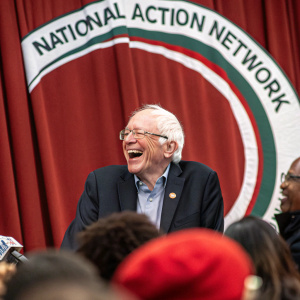 Former Senator Bernie Sanders laughing at a political rally. Behind him is a red curtain and the National Action Network logo.