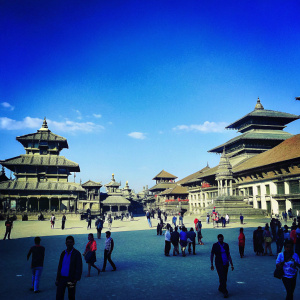 A vibrant photo capturing a bustling Patan Durbar Square in Kathmandu, Nepal. UNESCO World Heritage site with ongoing restoration. Diverse crowd of tourists and locals, clad in casual wear and some carrying backpacks. Ornate Newari architecture, detailed woodwork, and tiered rooftops. Clear blue sky overhead.