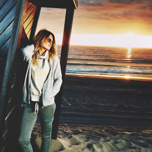 Maria Menounos, in a casual outfit, standing alone on a sandy beach at sunset.