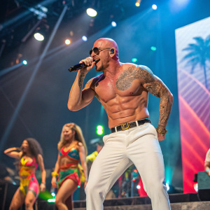 Pitbull, shirtless, performing on a brightly lit stage at a music festival. Tattooed arm visible. Surrounding by dancers in vibrant outfits.