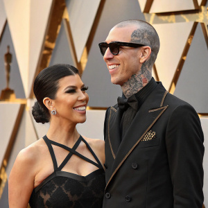 Kourtney Kardashian and Travis Barker, dressed up, smiling at Vanity Fair Oscar Party. Background is geometric shapes in gold and black.