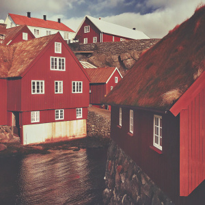 In the heart of Tórshavn, Faroe Islands, stands Tinganes, a charming village. It boasts vibrant red, white, and contrasting buildings with steep roofs. The town sits on a rocky coastline, reflected in the surrounding dark blue waters, beneath a sky filled with wispy clouds.