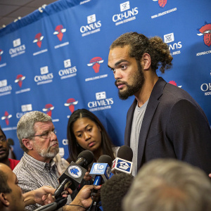 Joakim Noah during a press conference. Intense discussions with reporters. Serious expressions all around. Blue backdrop with company logos.