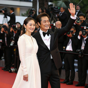 Kim Tae Hee and Lee Byung Hun, both smiling and waving at fans. Kim Tae Hee in a long white dress, Lee Byung Hun in a black tuxedo. Crowded red carpet with flashing cameras and paparazzi.