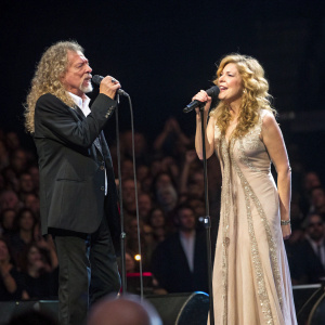 Robert Plant and Alison Krauss performing live at the O2 arena. Both are dressed in formal attire. Plant in black suit, Krauss in elegant floor-length gown. Crowd visible in background