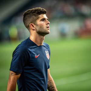 Christian Pulisic, male soccer player, intense expression. Green sports field background, blurred.