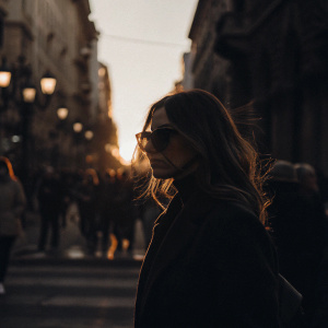 Monica Bellucci, with long wavy brown hair and dark sunglasses, walking down a bustling city street at sunset.
