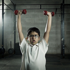 A young Asian boy, intense expression, short black hair, white polo shirt, lifting red dumbbells overhead. Gym background