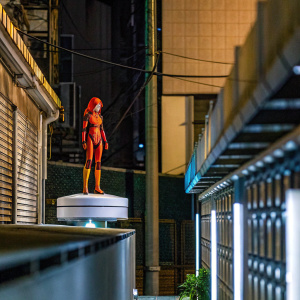 Digital 3D rendering of a figurine resembling Asuka Langley from Neon Genesis Evangelion. The character stands on a white, flat circular base against an orange backdrop. The figurine's attire and hairstyle are intricately detailed.