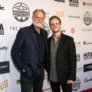 Taylor Hackford and Alex Hackford, two caucasian men, are at the Music Supervisor Awards gala. They both wear dark blazers and stand confidently in front of a stark white wall adorned with logos and sponsor names from various brands.