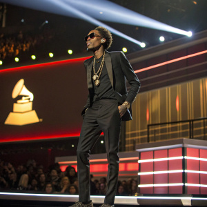 Wiz Khalifa at the Grammy Awards, sporting a sleek black suit and gold chains. Confident pose on stage with a spotlight