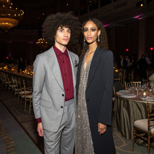 Hari Nef and Elaine Welteroth at a fashion gala. A young man with an afro hairstyle in a gray suit and maroon undershirt. They are in a spacious, dimly lit room with elegant decor.