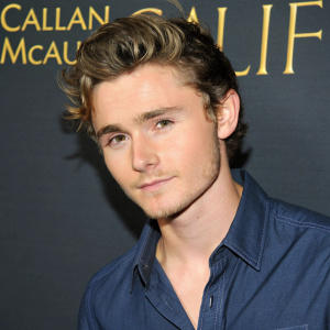 Close-up photo of Callan McAuliffe, a young actor with tousled hair and neat haircut, wearing a dark blue shirt. Shot against a plain, dark background with golden-yellow text. High-resolution image.