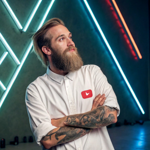 Felix Kjellberg, aka PewDiePie, sporting a large beard and tattoos, posing confidently. White shirt with YouTube logo. Neon light background with geometric shapes